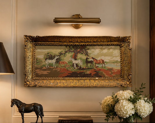 A framed needlepoint tapestry depicting horses in a field under a tree, hung on a beige wall below a brass wall sconce. The frame is ornate gold.