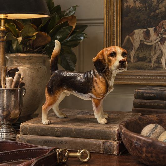 A realistic beagle dog figurine standing on stacked vintage books on a wooden table, surrounded by a lamp, potted plant, framed dog painting, books, and a leather accessory in a styled interior setting.