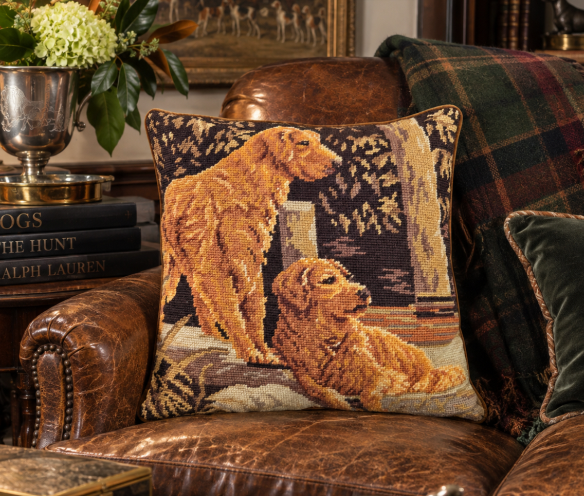 A square needlepoint throw pillow featuring two golden retriever dogs in a woodland scene, styled on a brown leather armchair next to a green plaid throw blanket.
