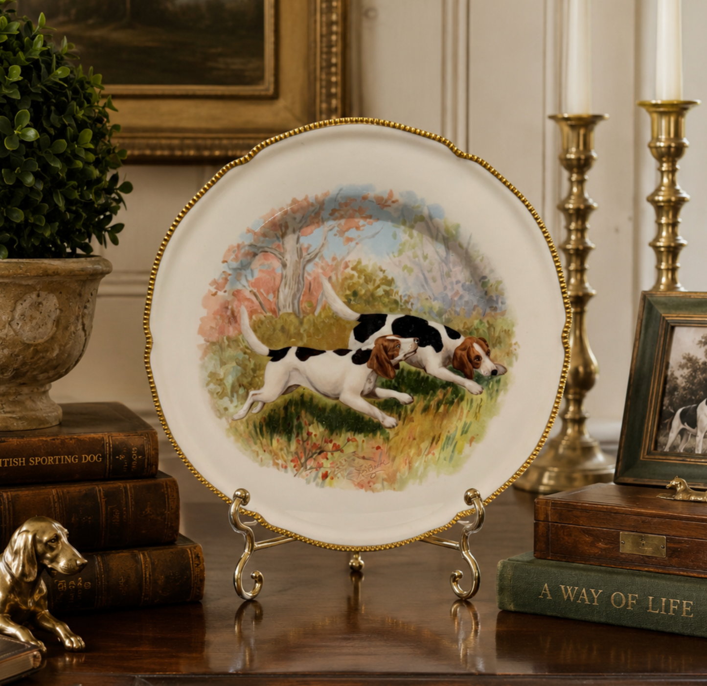 A decorative porcelain plate with a gold-trimmed rim and an illustration of two hunting dogs in a grassy field, displayed on a small metal stand on a wooden table in an antique-style room setting.