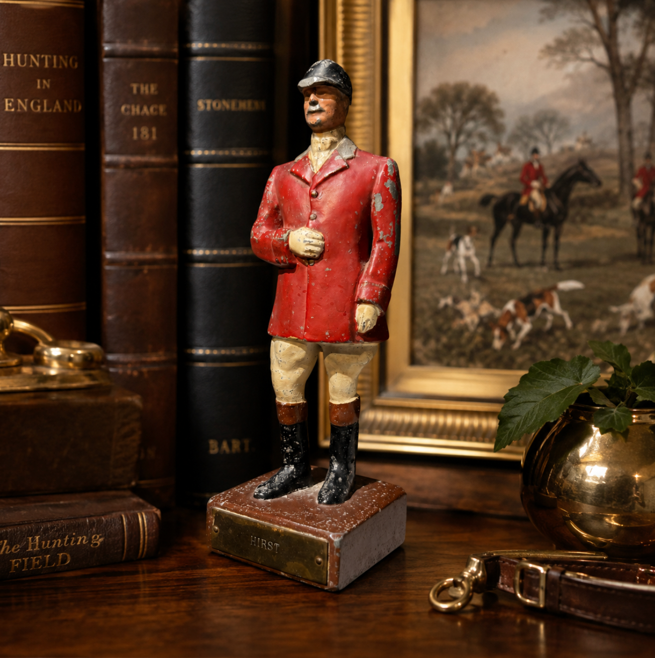 A vintage wooden hunting figurine of a foxhunter in a red jacket and beige trousers, standing on a small base labeled 'HIRST. The figurine is styled on a wooden table next to antique books, a brass pot, and a framed hunting painting.