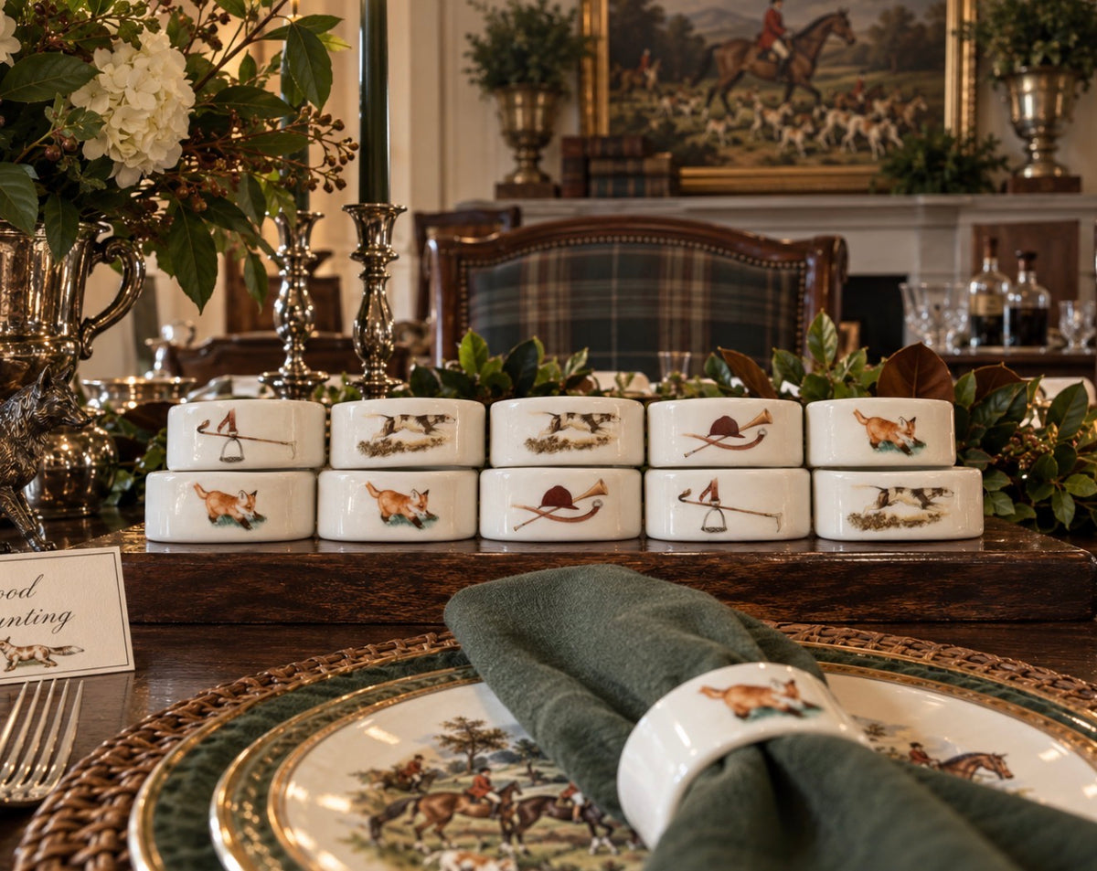 A set of white ceramic napkins with hand-painted hunting-themed illustrations, displayed on a wooden table runner in a styled dining room setting.
