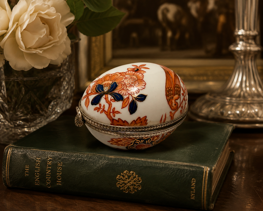 A decorative oval ceramic egg box with floral and bird motifs sits on a dark green vintage book, styled with a white rose in a glass vase and a silver candlestick in a warm indoor setting.