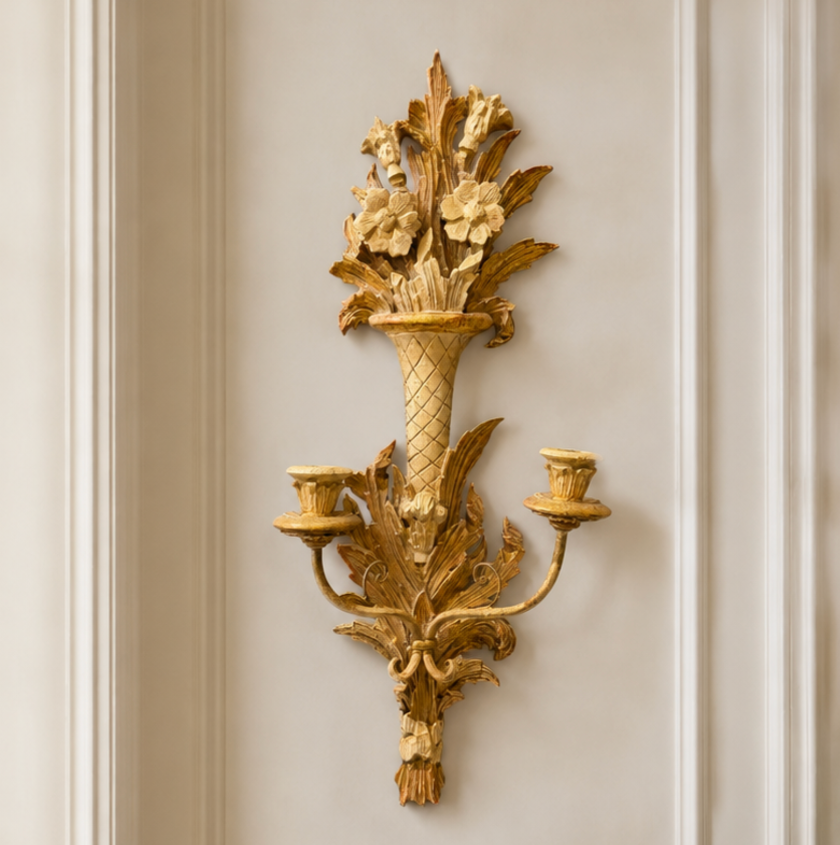 An ornate gold wall sconce featuring a sculpted vase filled with acanthus leaves and flowers, mounted on a cream paneled wall.
