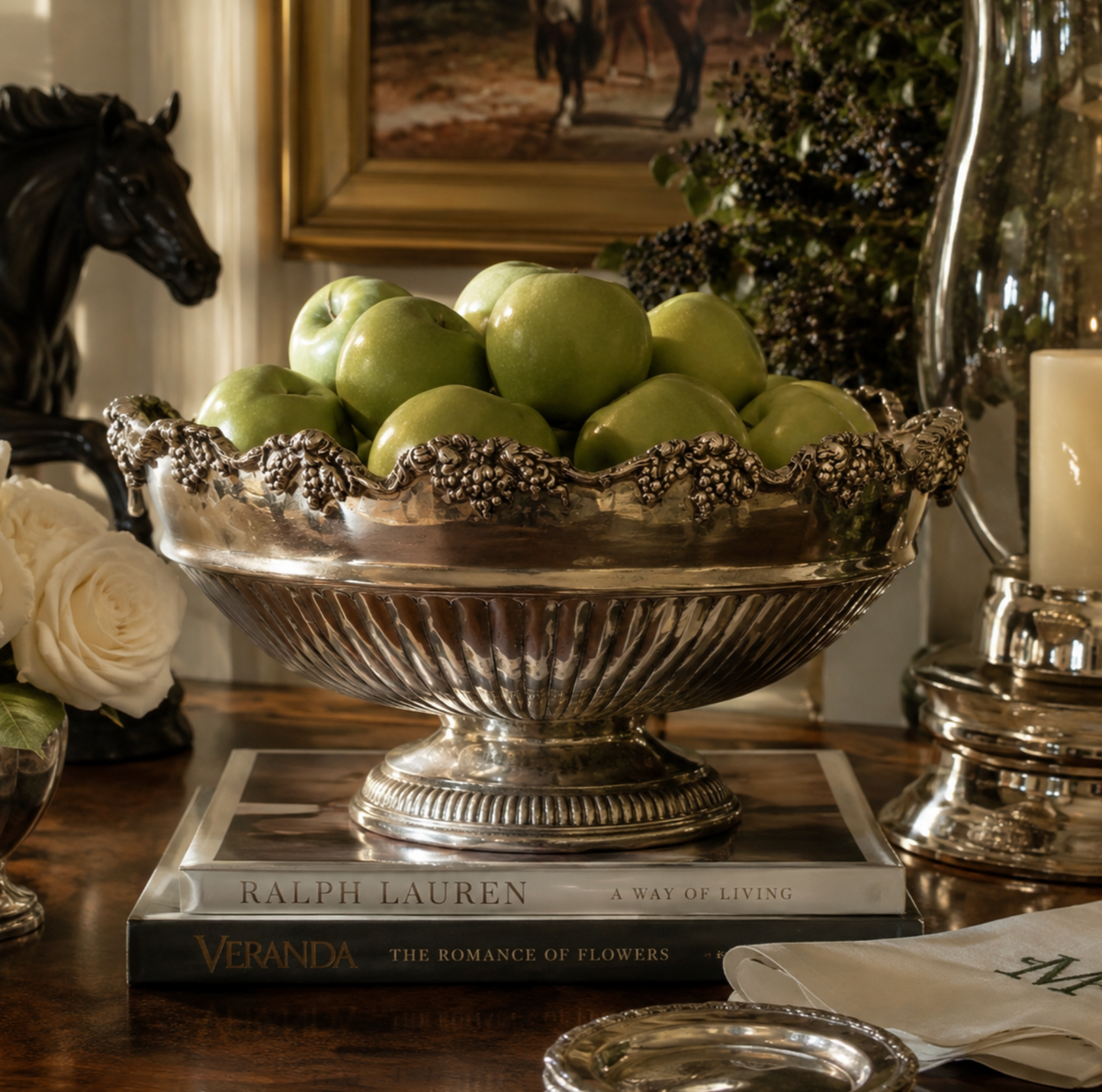 A large ornate silver pedestal bowl filled with green apples, placed on a stack of books on a wooden table in a styled interior setting.