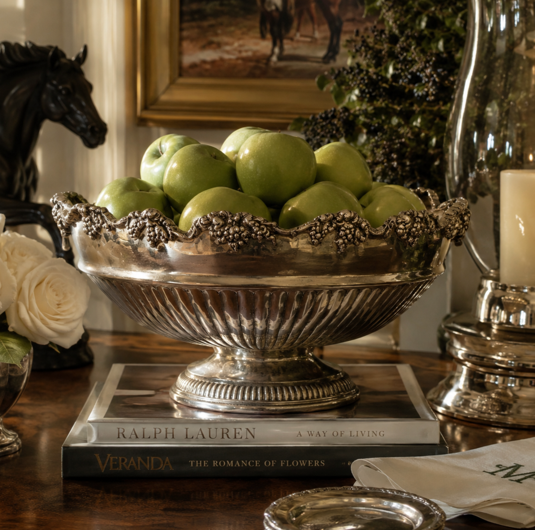 A large ornate silver pedestal bowl filled with green apples, placed on a stack of books on a wooden table in a styled interior setting.