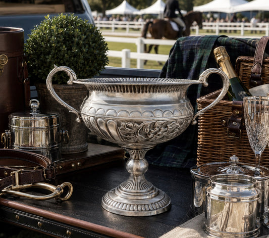 An ornate silver pedestal urn with decorative floral relief and looped handles, displayed on a dark trunk alongside leather accessories, a wicker basket, a champagne bottle, and a crystal glass. In the blurred background, a horse and rider and white event tents are visible outdoors.