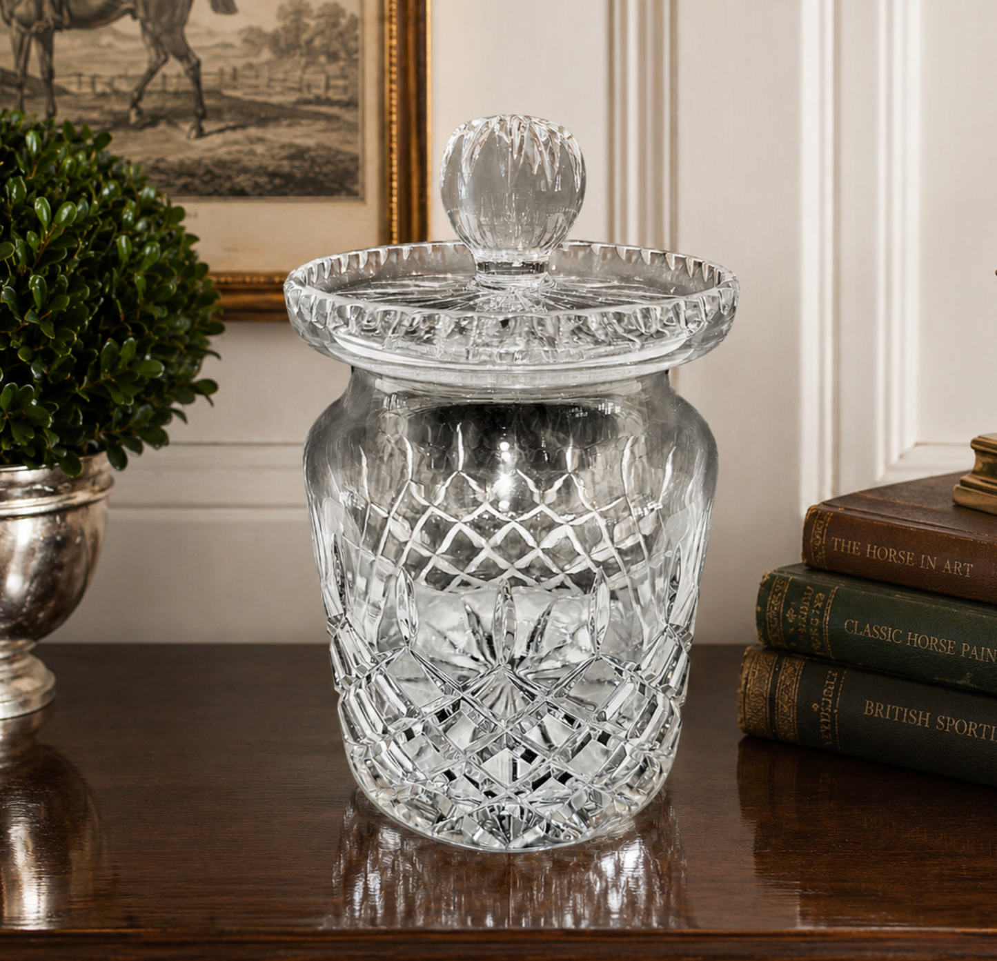 A clear cut-glass jar with a matching glass lid and knob, displayed on a dark wooden table in a styled interior setting with a potted plant and books.