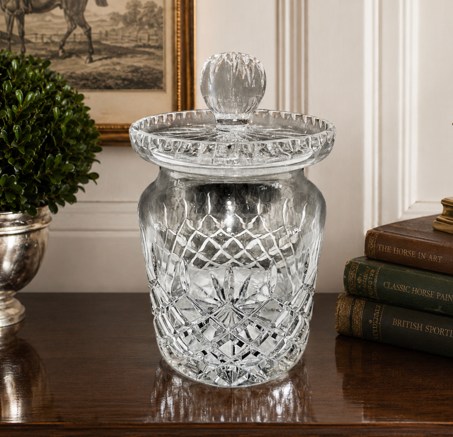 A clear cut-glass jar with a matching glass lid and knob, displayed on a dark wooden table in a styled interior setting with a potted plant and books.