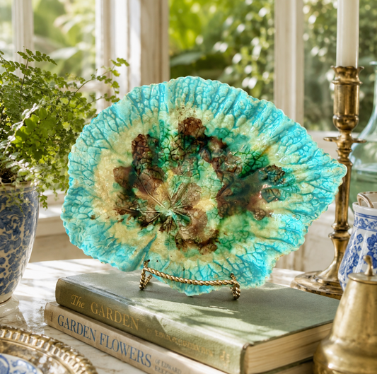 Decorative plate with a floral design on a stand, placed on books with a window and plants in the background.