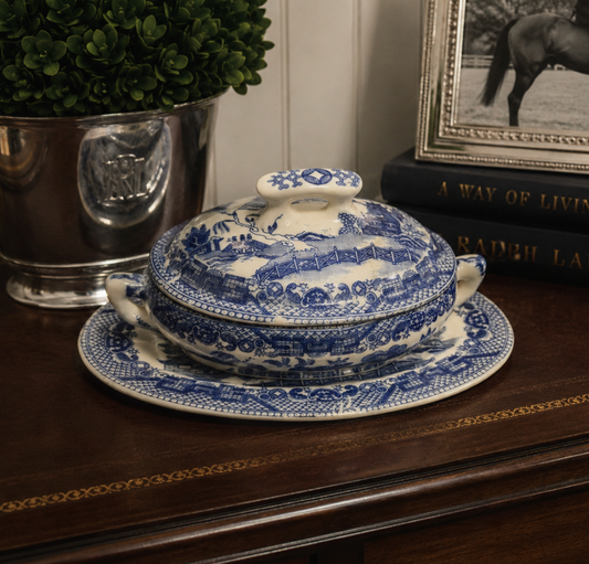 A blue and white ceramic lidded serving dish with handles, set on a matching saucer on a dark wooden table, styled with a silver planter and framed picture in the background.