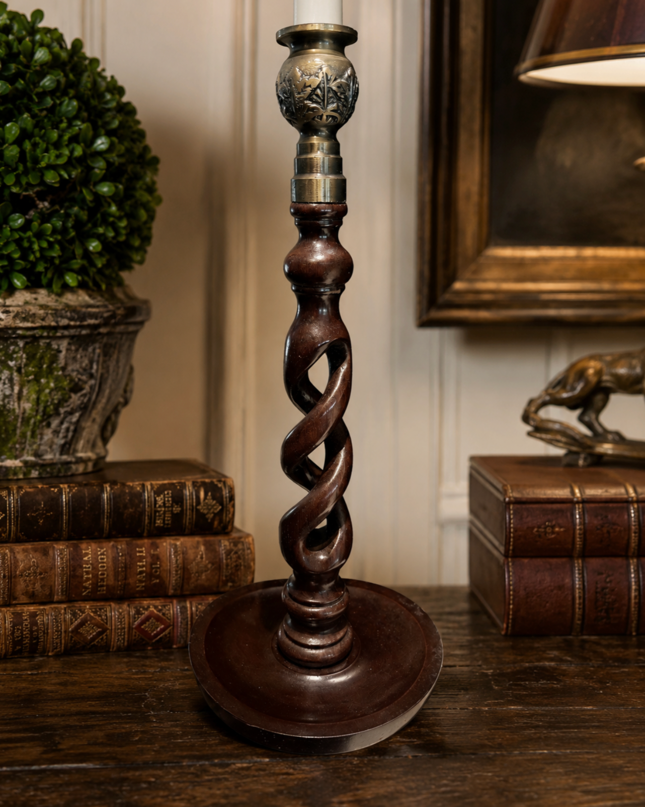 A dark brown twisted column candlestick or lamp base with ornate metallic accents near the top, displayed on a wooden table in a styled interior setting with books and a lamp in the background.