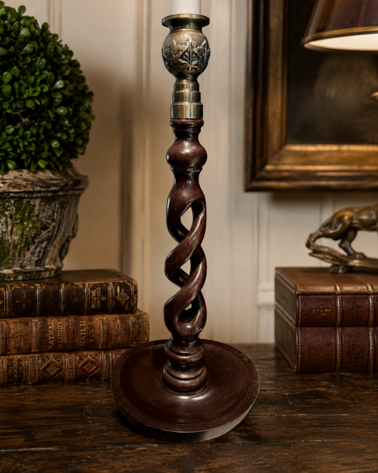 A dark brown twisted column candlestick or lamp base with ornate metallic accents near the top, displayed on a wooden table in a styled interior setting with books and a lamp in the background.