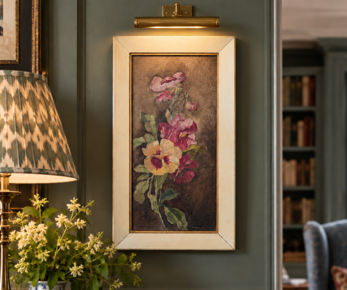 A framed vintage-style floral painting hangs on a dark green paneled wall in a cozy room setting, with a lamp, flowers, and a bookshelf visible.