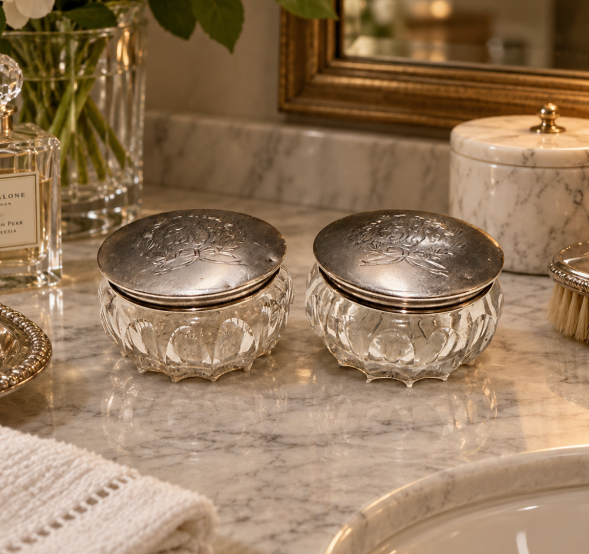 Two ornate clear glass jars with embossed floral lids and silver-toned metal rims, displayed on a polished marble bathroom counter.
