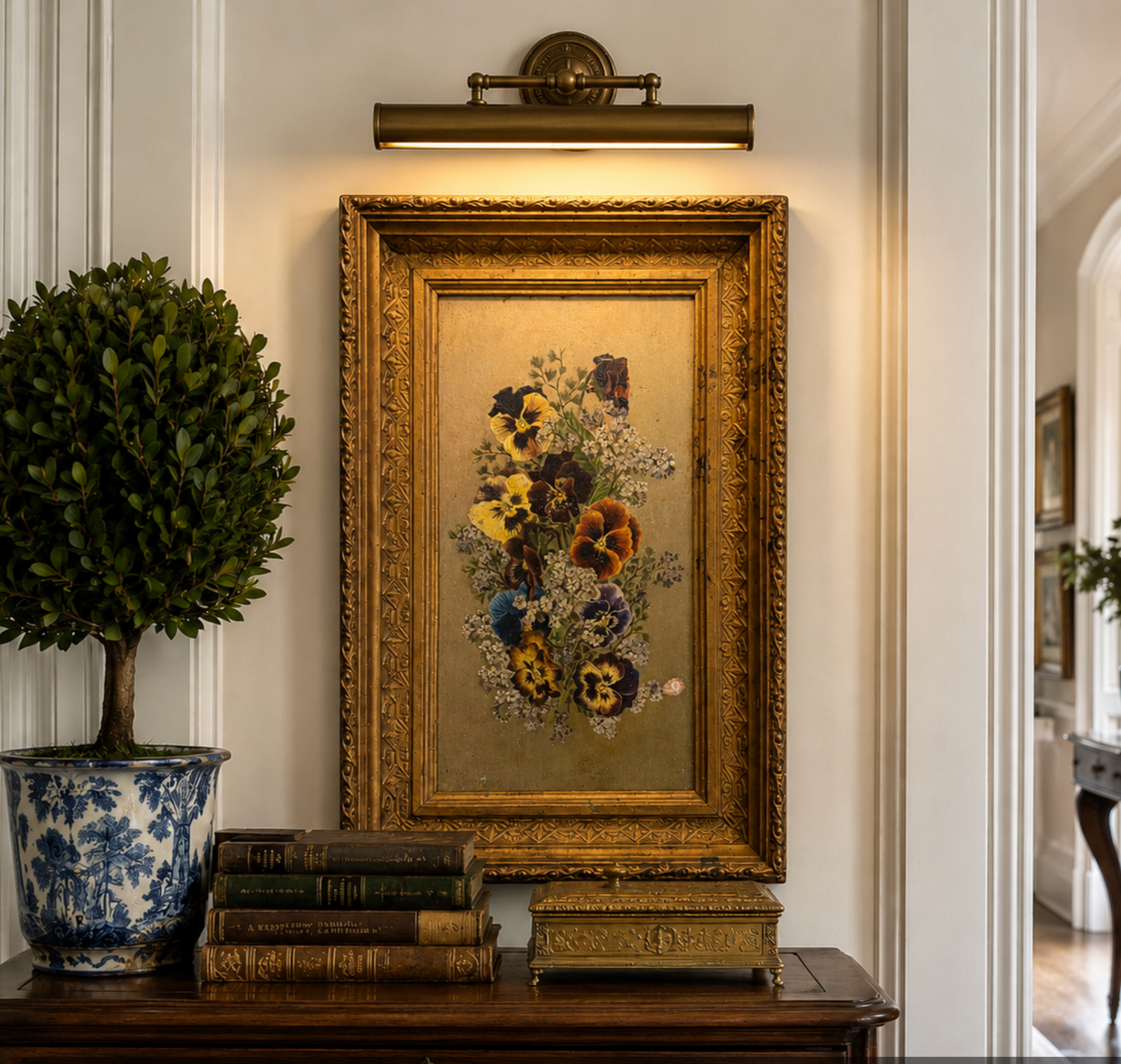 A framed vintage floral painting with a distressed gold frame hangs on a white paneled wall above a dark wooden console table. The scene includes a potted topiary tree in a blue-and-white ceramic pot, stacked antique books, and a small ornate box.