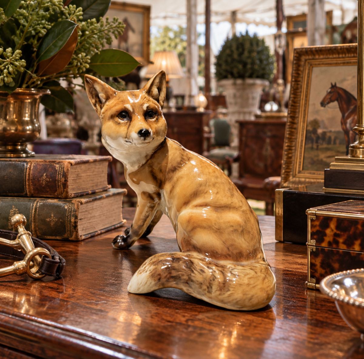 A glossy ceramic fox figurine in tan, cream, and brown tones, posed on a polished wooden table in an antique shop setting with books, framed art, and decor in the background.