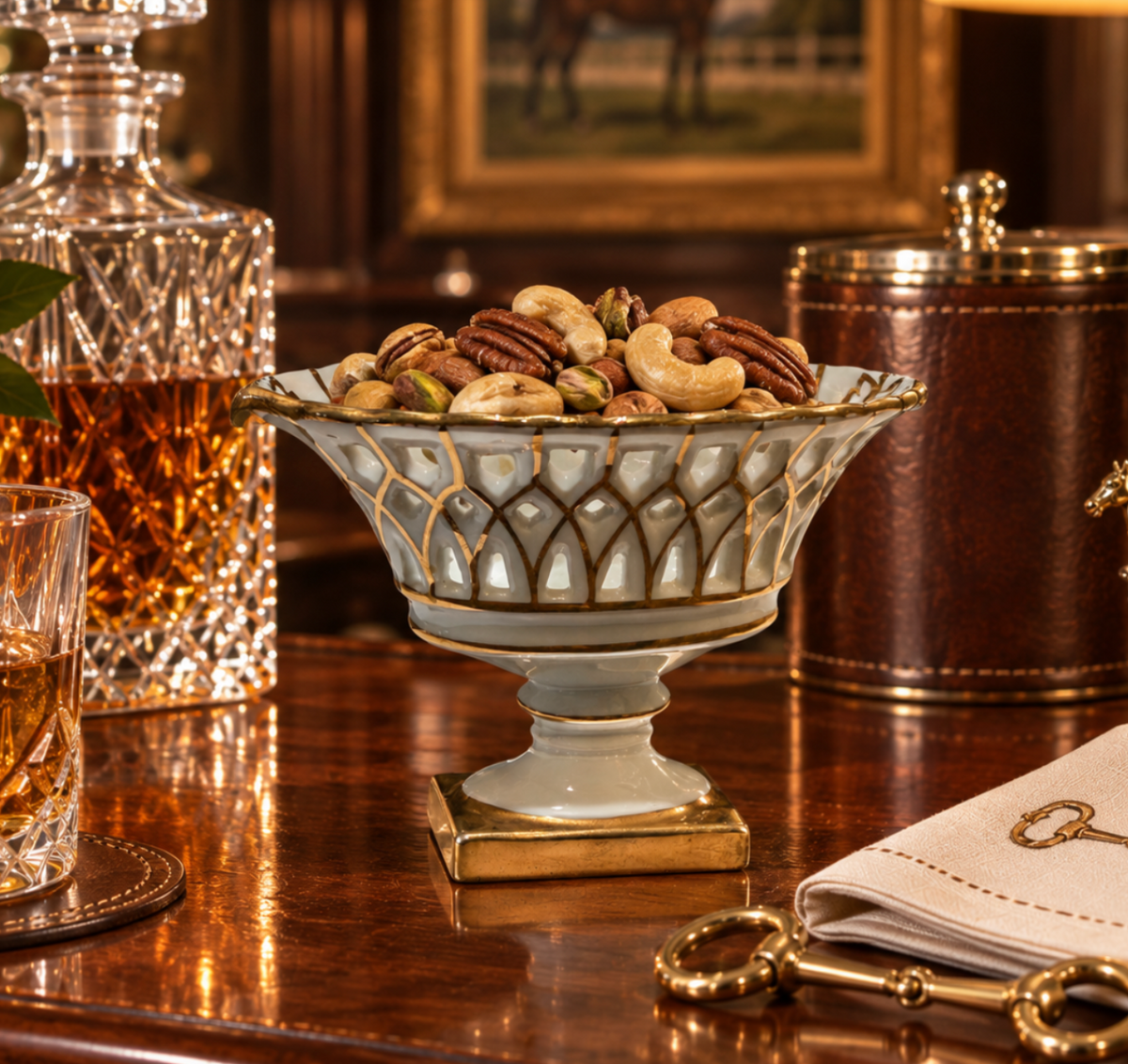 An ornate pedestal bowl with a white glaze and gold trim, filled with nuts and pecans on a polished wooden table, with a decanter, glass, and leather container in the background.