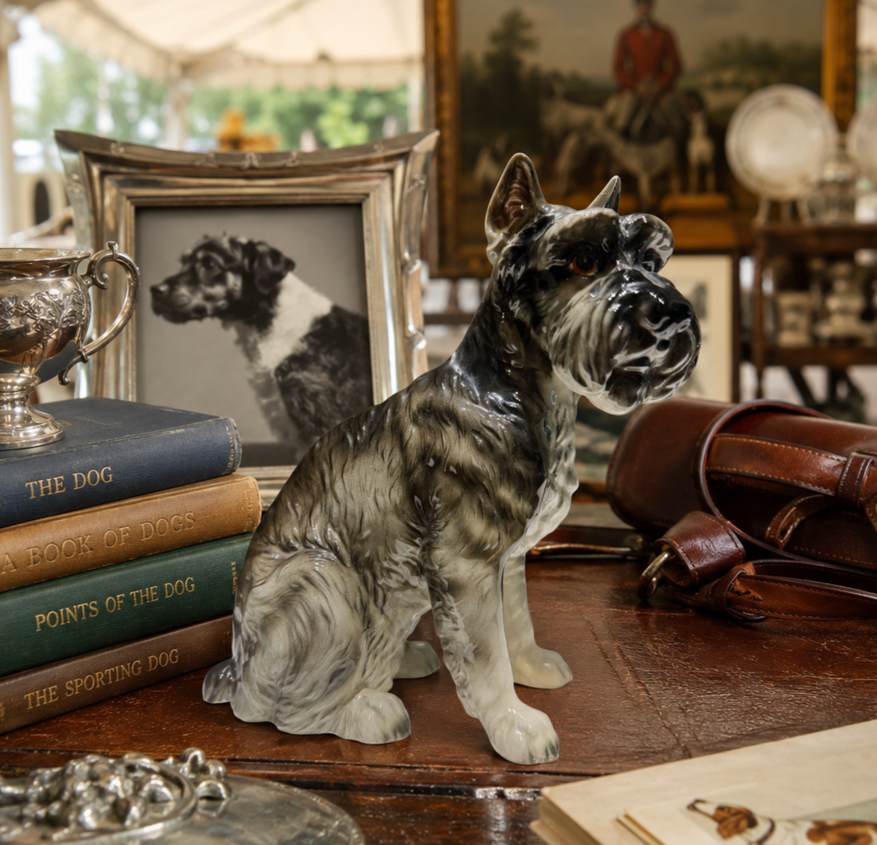 A ceramic figurine of a black and white Schnauzer dog sitting on a wooden table, surrounded by vintage books, a leather bag, a silver cup, and a framed picture of a dog.