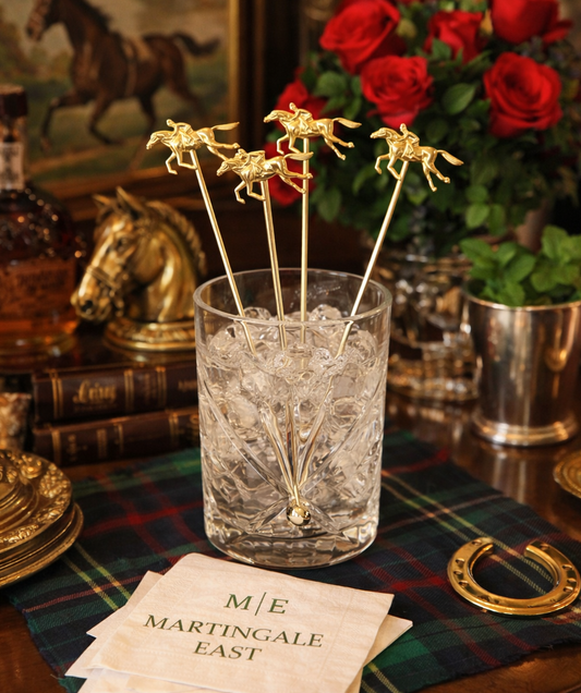 Glass with ice and gold stir sticks on a table with decorative items and red flowers.