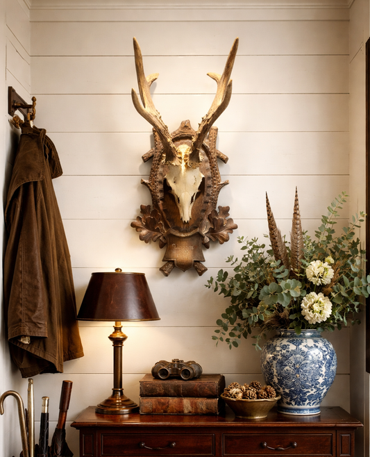 Decorative interior with deer skull, lamp, books, and flowers on a wooden console against a white paneled wall.