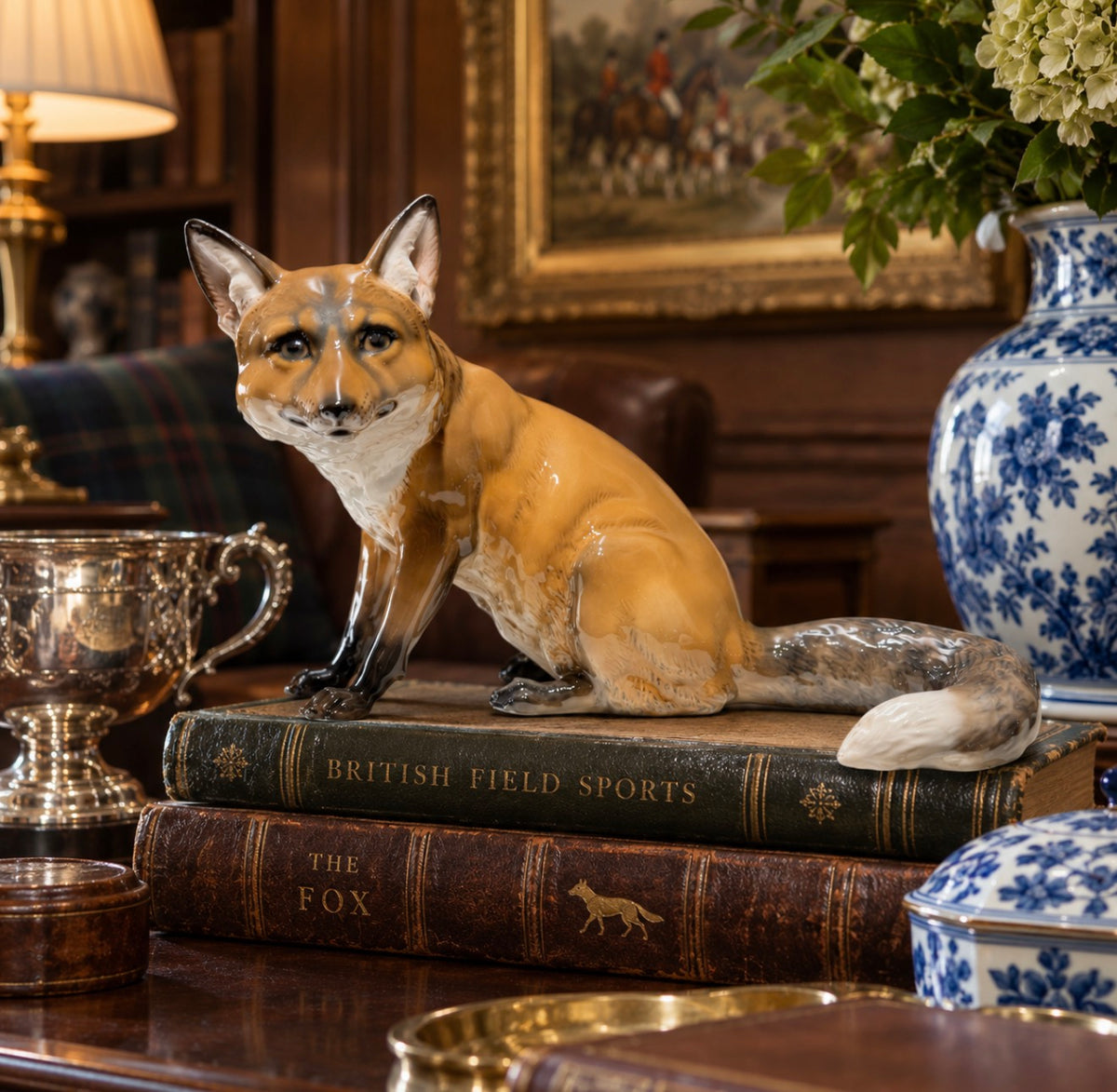 A ceramic fox figurine in a sitting pose, displayed on top of two vintage-style books on a wooden table in a classic, warm-toned room setting.