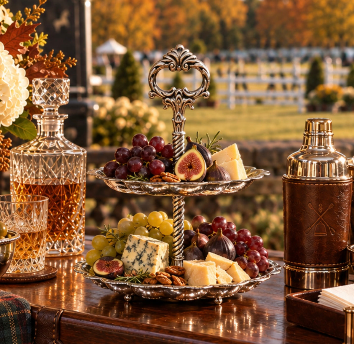 An ornate two-tiered silver serving stand filled with grapes, figs, cheese, and nuts on a wooden table, with a cut glass decanter and a leather-and-gold cocktail shaker nearby in an outdoor garden setting.