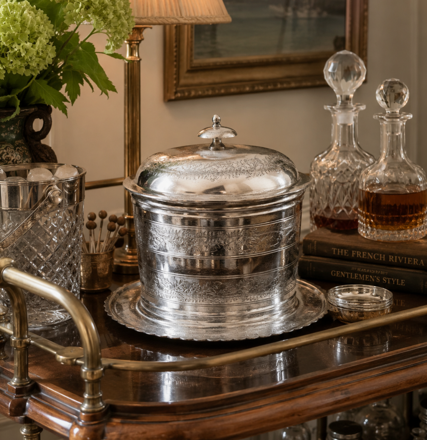 A polished silver lidded canister or tumbler with engraved detailing sits on a dark wooden bar cart, surrounded by glass decanters, books, and a vase in an elegant indoor setting.