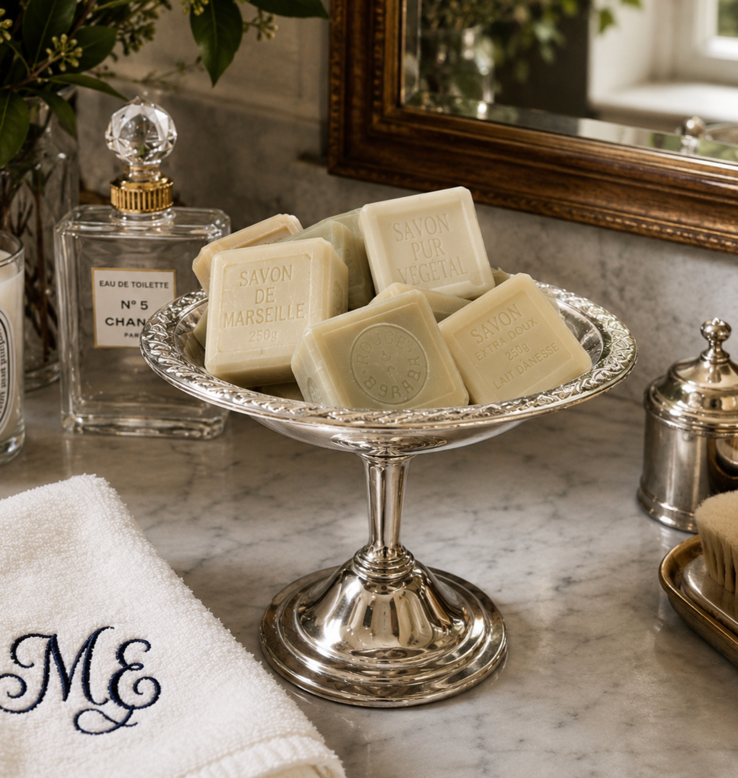 Several rectangular bars of Savon de Marseille soap in off-white cream are arranged on an ornate silver pedestal stand on a marble bathroom counter. A white monogrammed towel, a perfume bottle, a glass jar, and a mirror are visible in the styled setting.