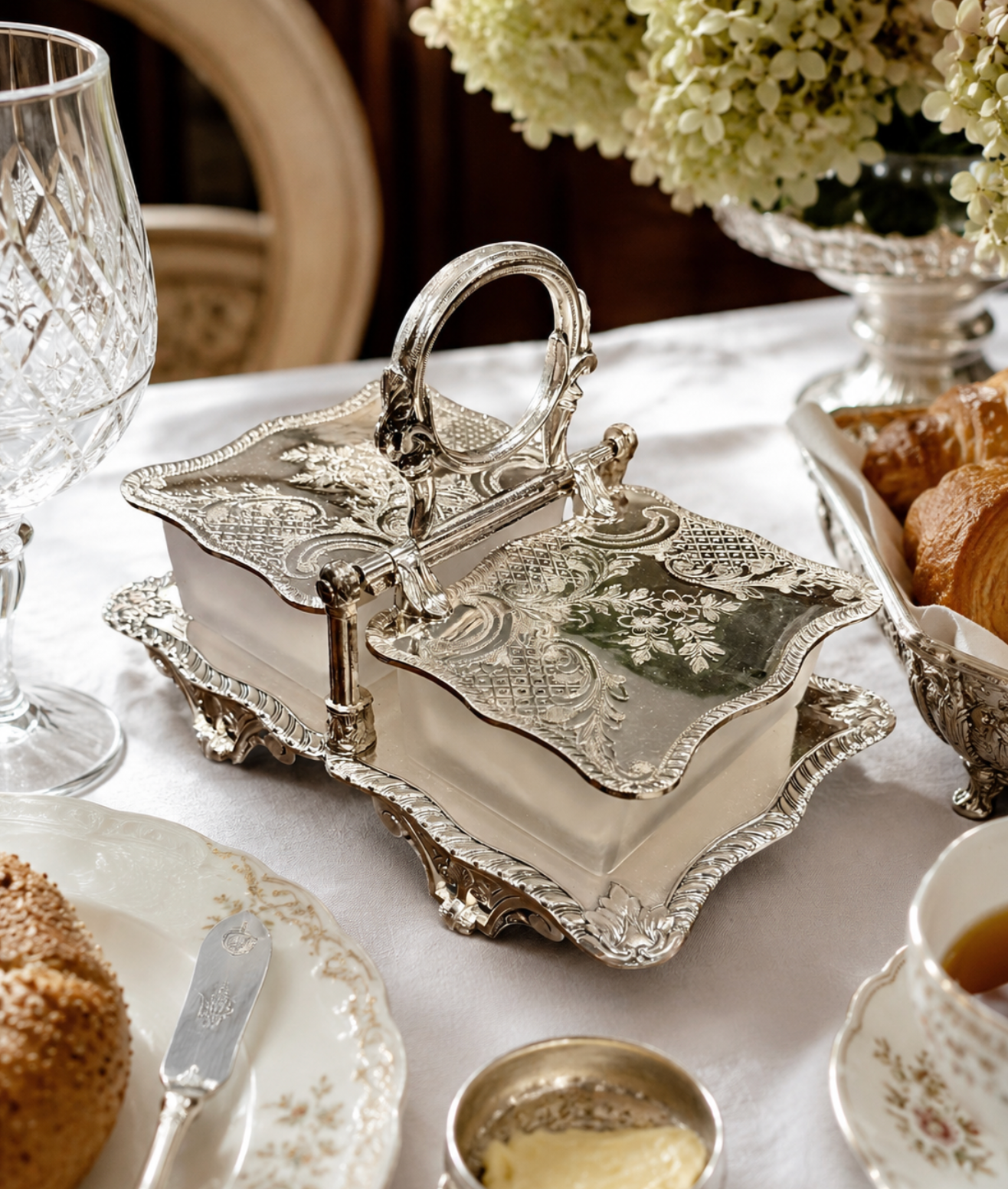An ornate silver-toned sugar bowl and creamer set with a matching handle, displayed on a white tablecloth in a styled breakfast setting with croissants, a glass, and floral details.