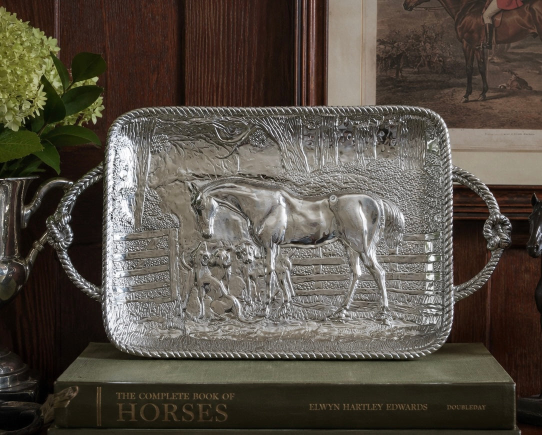 A rectangular silver-toned decorative tray with rope-style handles, featuring an embossed relief of a horse and rider scene, displayed on a book in a rustic indoor setting.