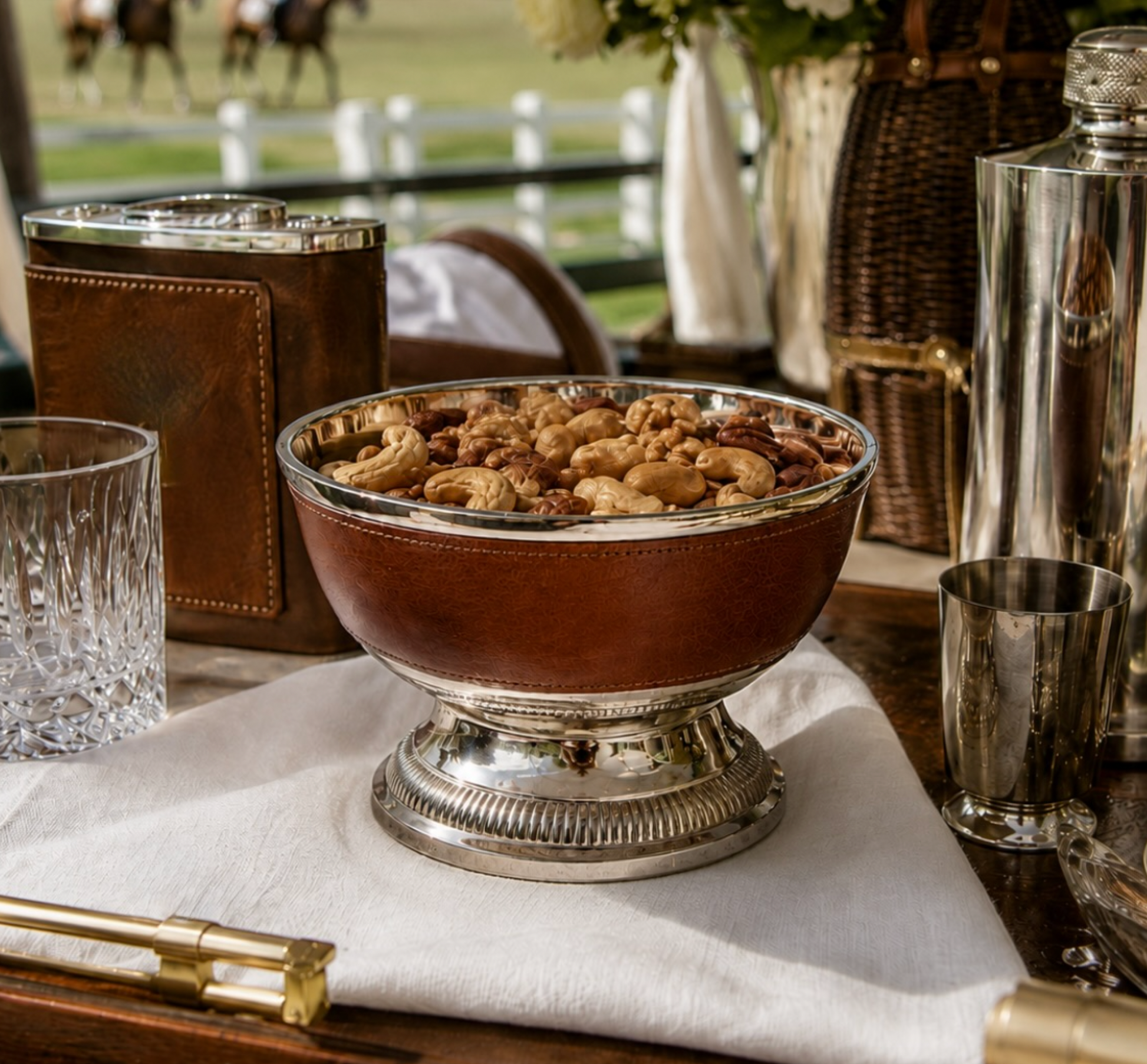 A brown leather-covered pedestal bowl filled with cashews and nuts, set on a white napkin on a wooden table with silver and brown accessories in a rustic outdoor setting.