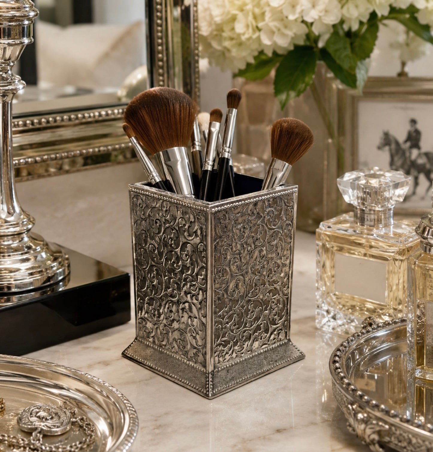 An ornate silver makeup brush holder with intricate floral embossing, filled with makeup brushes, set on a marble vanity table next to perfume bottles and a mirror.