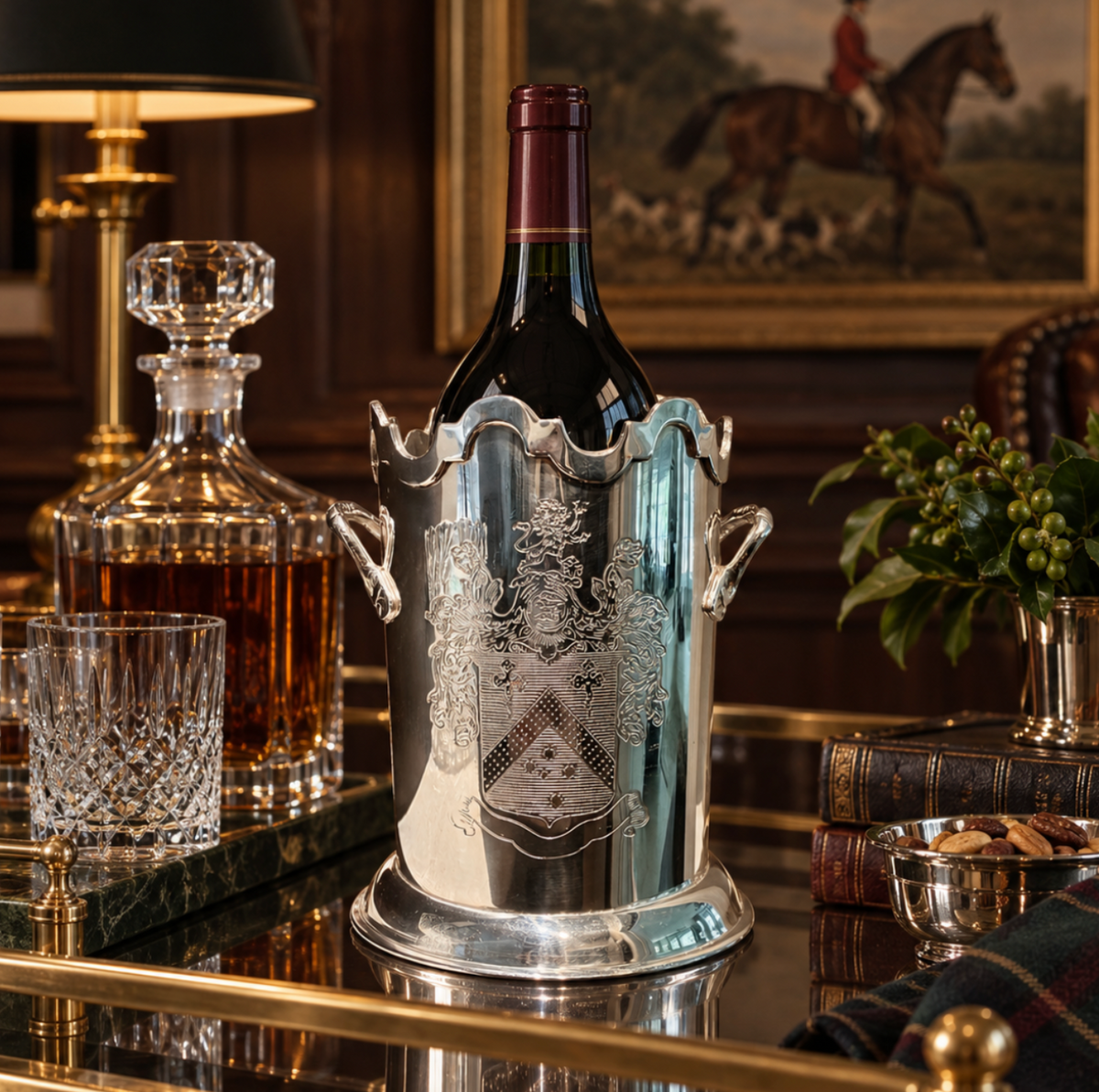 A polished silver wine bottle cooler bucket holding a dark wine bottle, set on a reflective table in an elegant room with a glass decanter, cut glass, and framed artwork in the background.