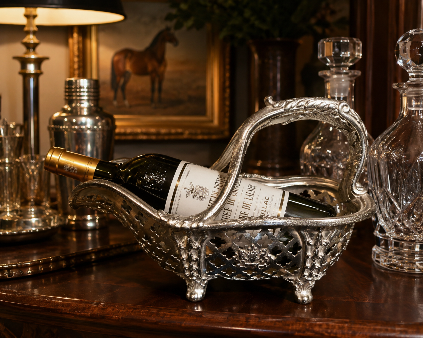 A silver ornate wine basket with legs and a curved handle, holding a wine bottle on a dark wooden table, surrounded by glass decanters and a lamp in a warm, elegant interior setting.