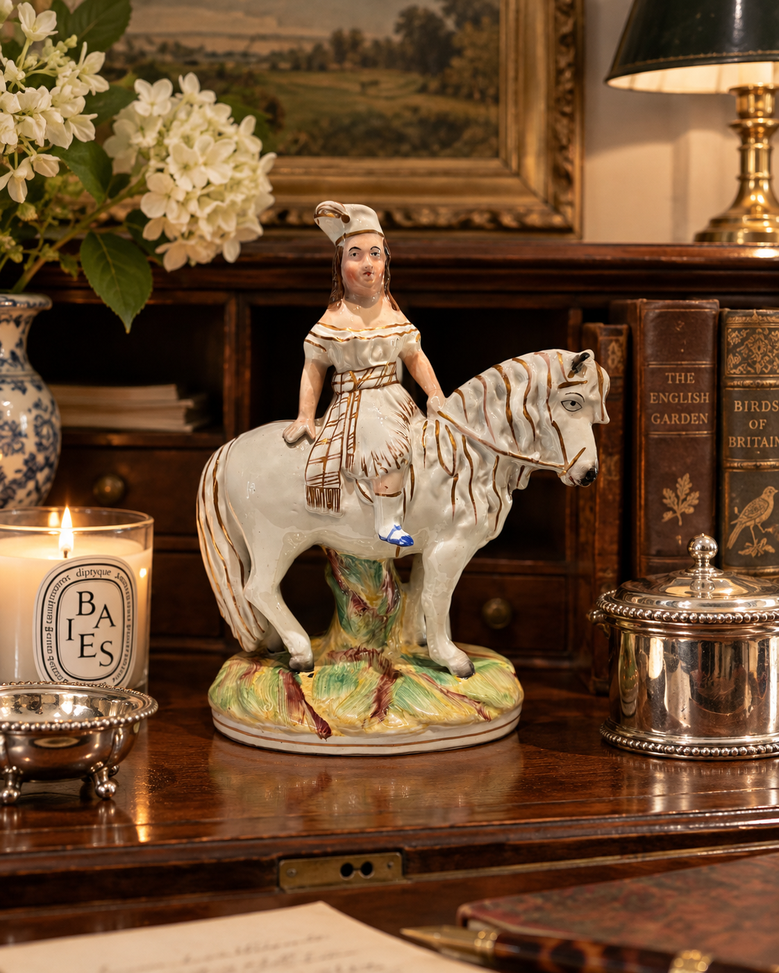 A ceramic figurine of a woman riding a white horse, displayed on a polished dark wood desk surrounded by decorative items including a lit candle, books, and a lamp.