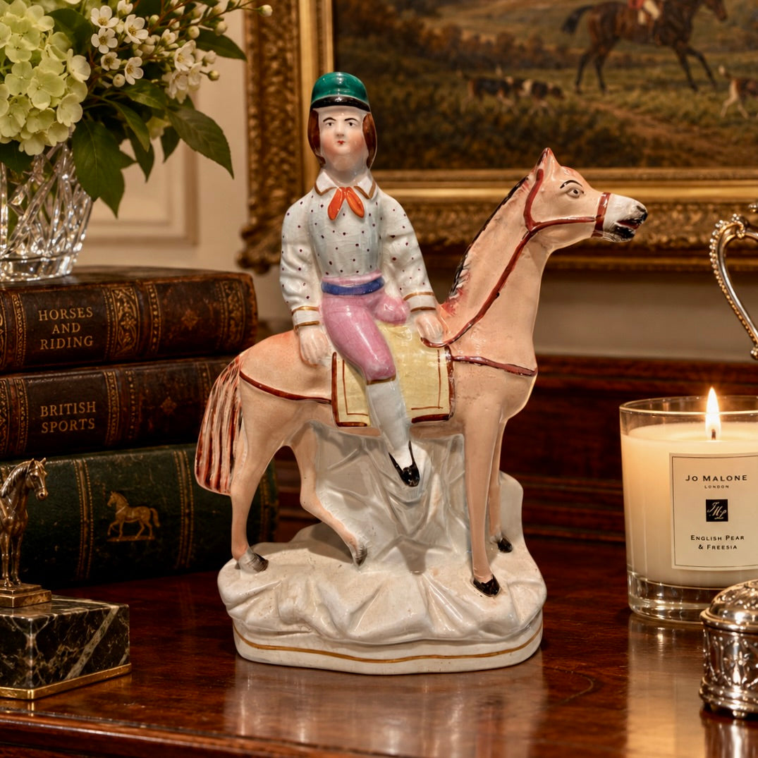 A ceramic equestrian figurine of a rider on a horse, displayed on a wooden table next to a lit Jo Malone candle, vintage books, and a glass vase of flowers.