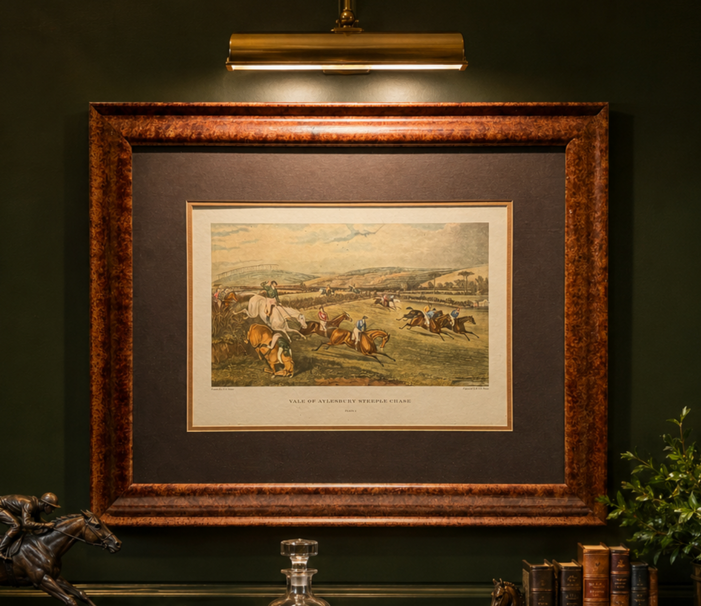 A framed vintage print depicting a horse race scene, mounted on a dark green wall under a brass wall lamp. The print features a detailed illustration of horses and jockeys in a field, with a wide brown mat and a textured bronze-brown wooden frame.