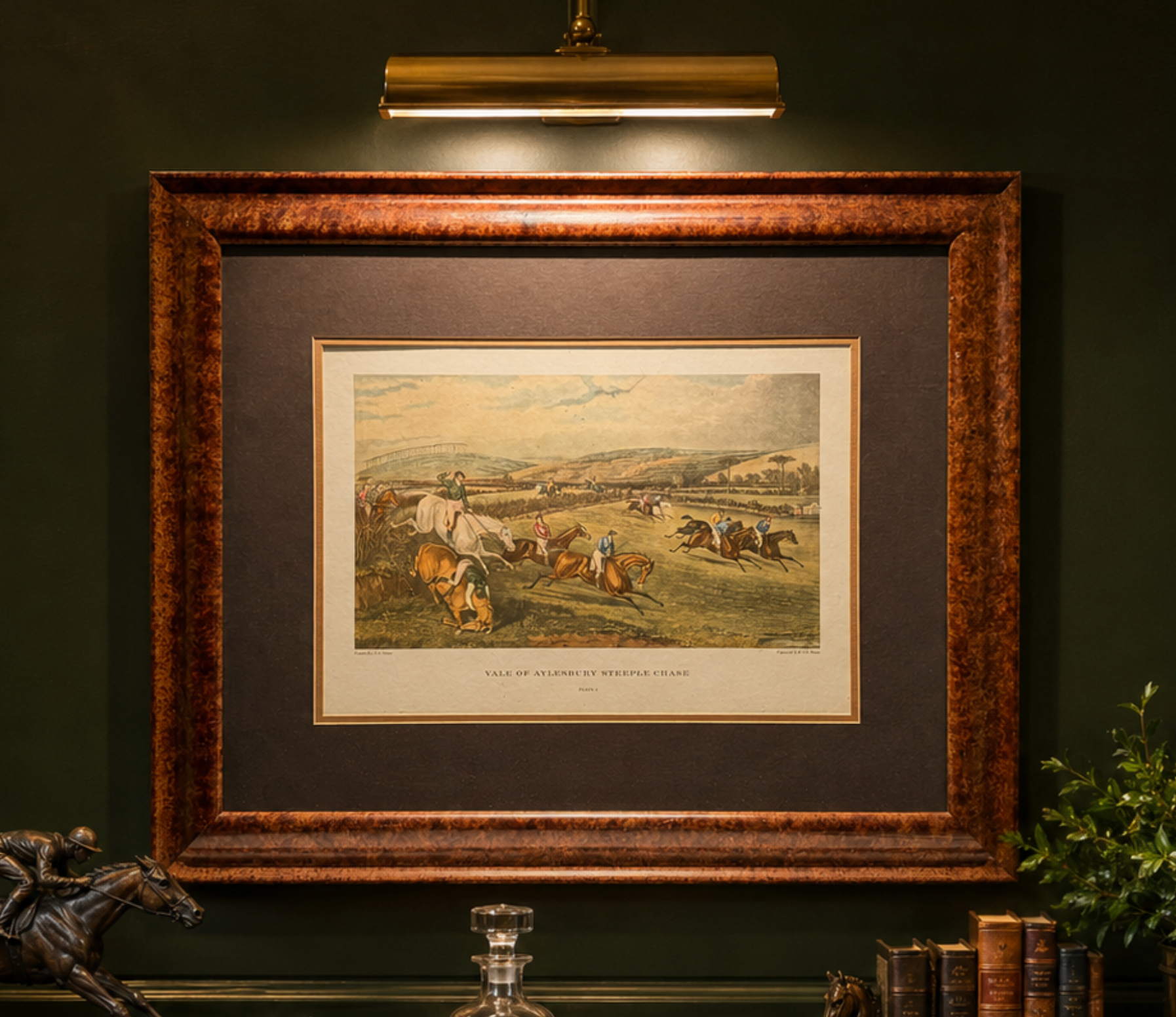 A framed vintage print depicting a horse race scene, mounted on a dark green wall under a brass wall lamp. The print features a detailed illustration of horses and jockeys in a field, with a wide brown mat and a textured bronze-brown wooden frame.