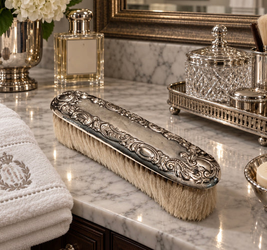 An ornate silver metal hair-brush brush with cream-colored bristles, resting on a white and gray marble bathroom vanity next to a folded white towel.