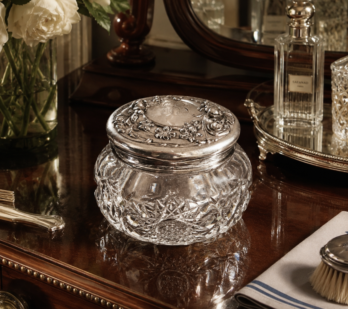 A clear cut-glass jar with a silver-toned ornate lid, displayed on a polished dark wood vanity table in a styled interior setting.
