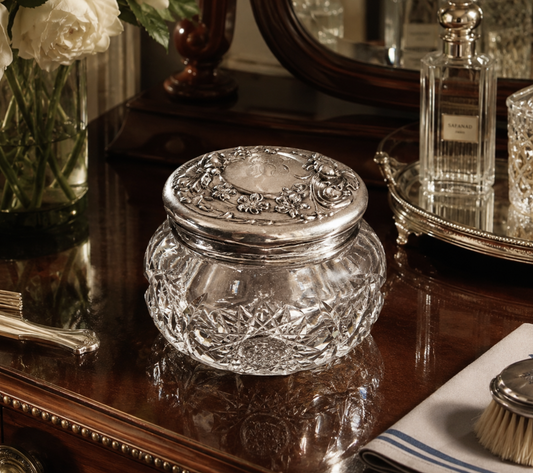 A clear cut-glass jar with a silver-toned ornate lid, displayed on a polished dark wood vanity table in a styled interior setting.
