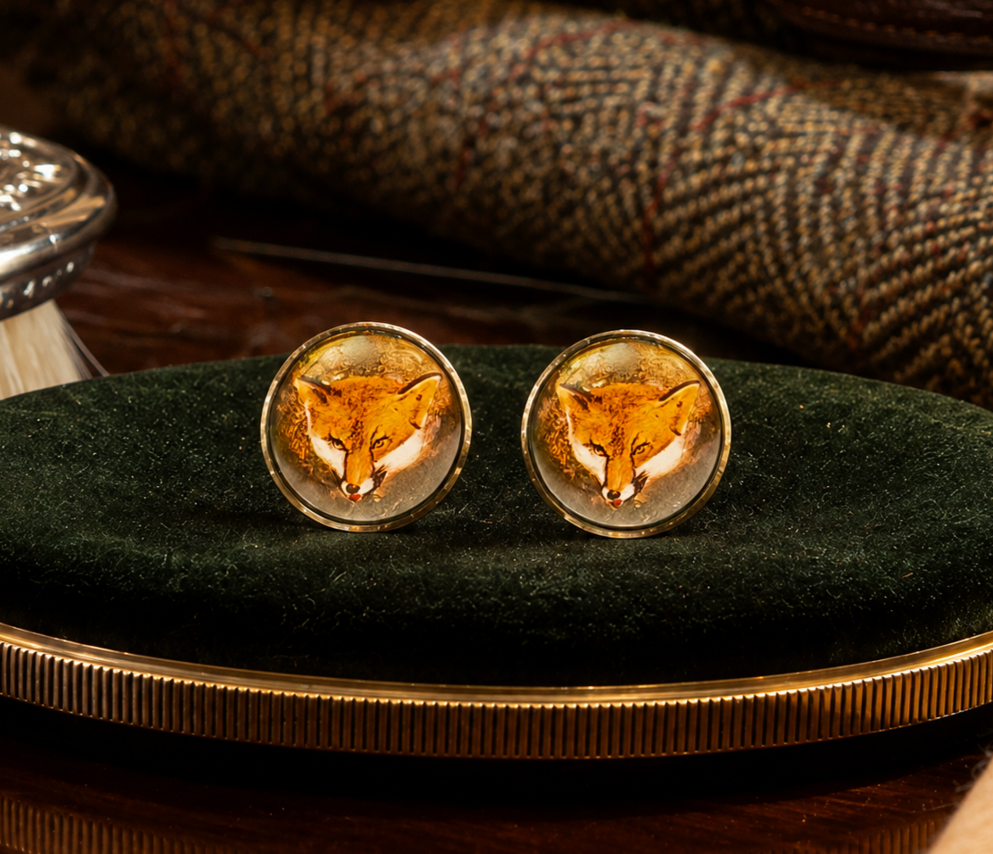 A pair of oval stud earrings with a detailed fox face design under clear resin, set in gold-tone metal, displayed on a dark green velvet cushion.