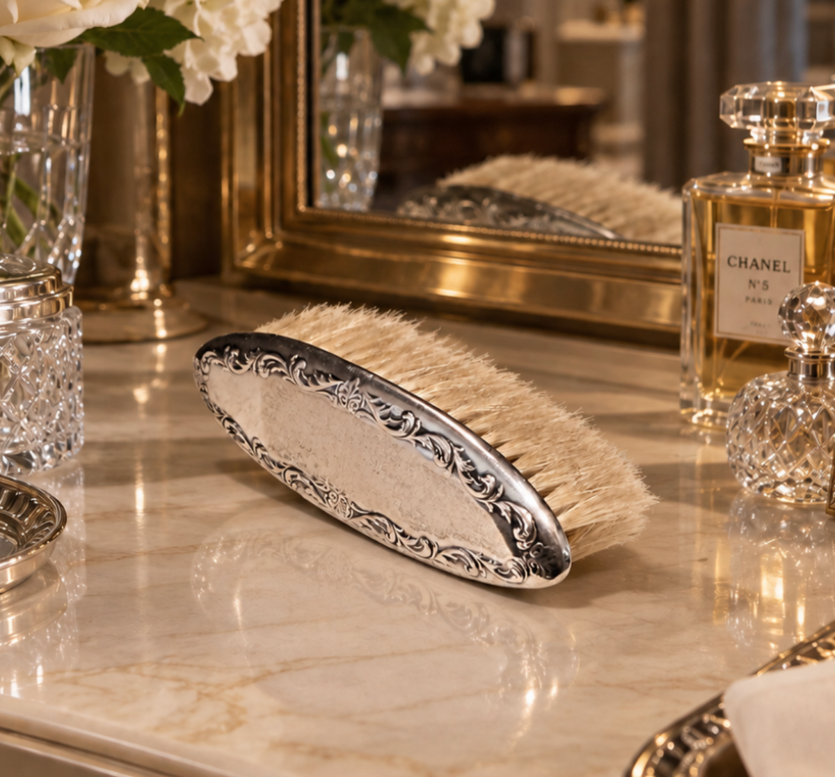 An ornate silver-handled hair brush with cream bristles, resting on a polished marble surface in a luxurious vanity setting with a Chanel perfume bottle and a mirror in the background.