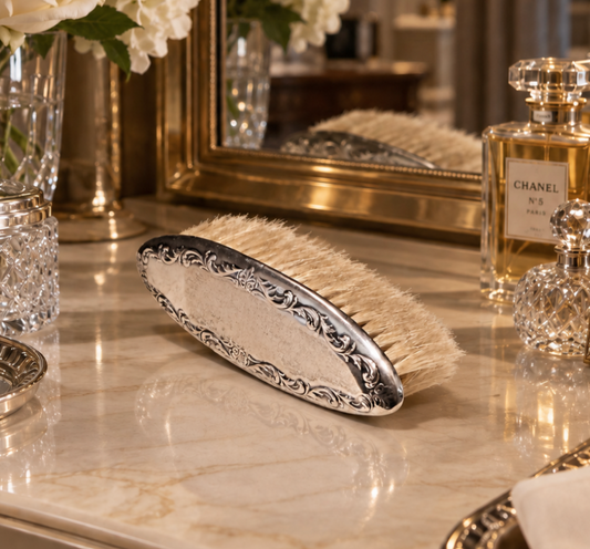 An ornate silver-handled hair brush with cream bristles, resting on a polished marble surface in a luxurious vanity setting with a Chanel perfume bottle and a mirror in the background.