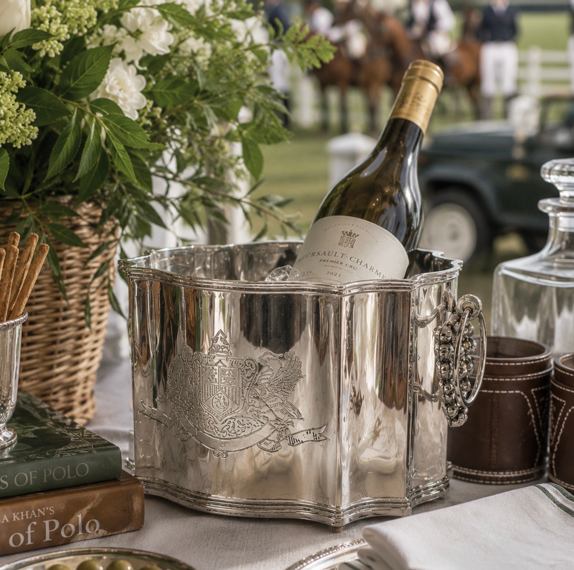 A polished silver wine cooler bucket holding a bottle of Brailley Champagne, styled on a table with a wicker basket of white flowers, books, and leather cups in an outdoor equestrian setting.