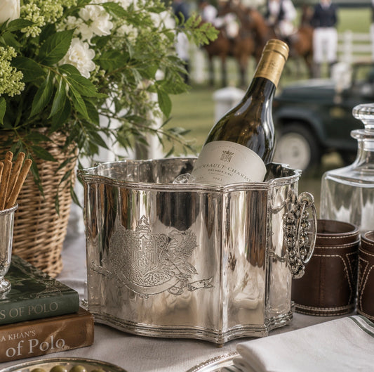 A polished silver wine cooler bucket holding a bottle of Brailley Champagne, styled on a table with a wicker basket of white flowers, books, and leather cups in an outdoor equestrian setting.