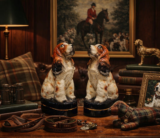 Two ceramic dog figurines on a wooden table with a painting and books in the background.