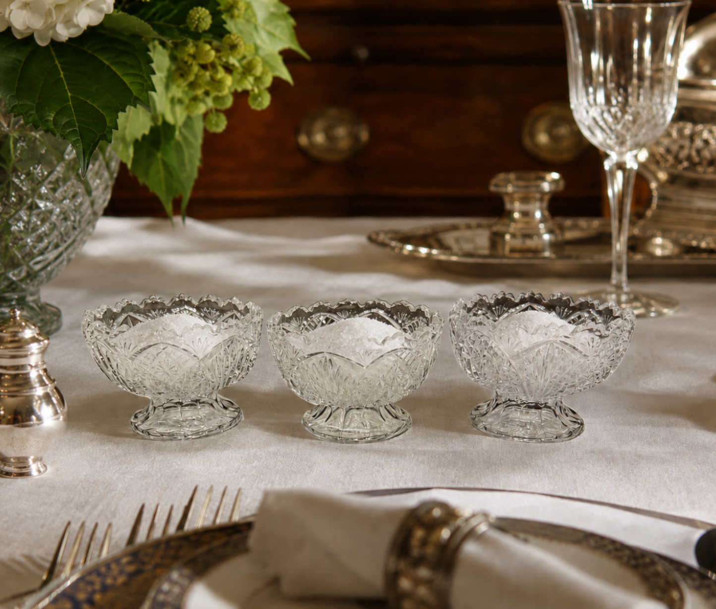 Three clear cut-crystal footed dessert or candy bowls arranged on a white tablecloth in a styled dining setting, with plates, a wine glass, flowers, and silver-toned tableware visible.