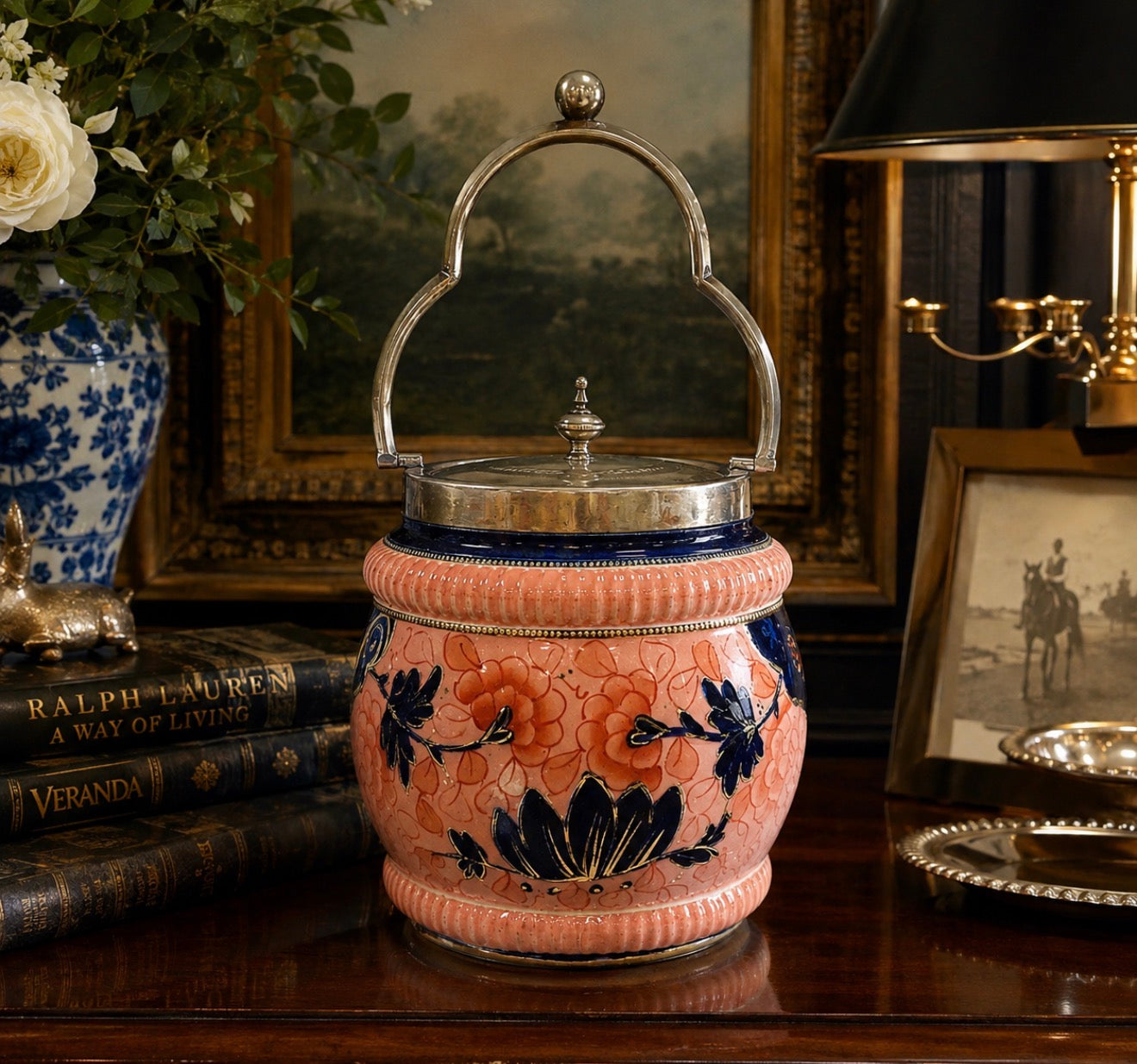 A decorative ceramic lidded jar or canister with a pink peach body, navy blue floral motifs, and a silver metal handle, displayed on a wooden table in an ornate room setting.
